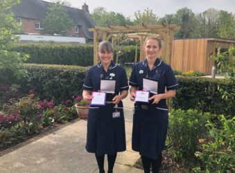 2 female nurses wearing dark blue uniform holding awards and smiling at the camera in Hospice garden