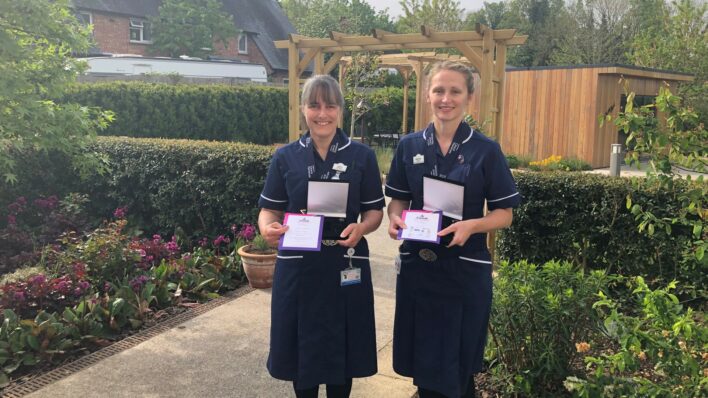 2 female nurses wearing dark blue uniform holding awards and smiling at the camera in Hospice garden