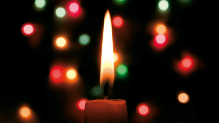 Candle at night with green red and yellow Christmas lights behind it