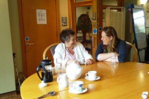 2 females sitting at a table with a teapot and cups, smiling at each other 