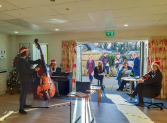 Group of Day Therapy Team colleagues singing accompanied by others on double bass, violin and guitar - in front of laptop at virtual Christmas party