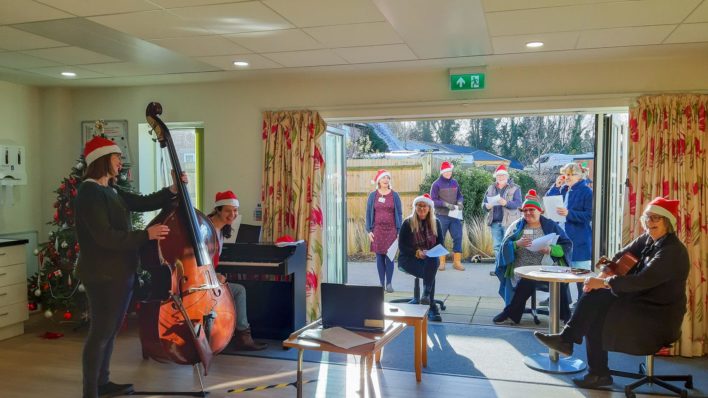 Group of Day Therapy Team colleagues singing accompanied by others on double bass, violin and guitar - in front of laptop at virtual Christmas party