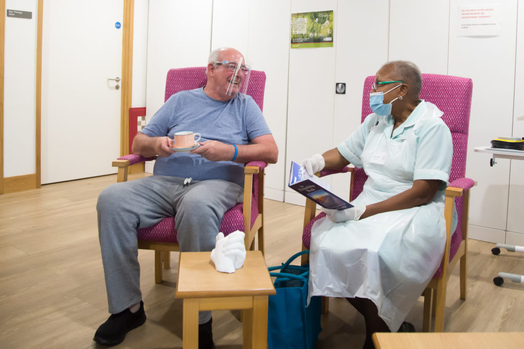 Patient wearing a perspex visor in day therapy enjoys a cup of tea, as he chats to a nurse wearing a face mask