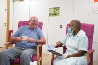 Patient wearing a perspex visor in day therapy enjoys a cup of tea, as he chats to a nurse wearing a face mask