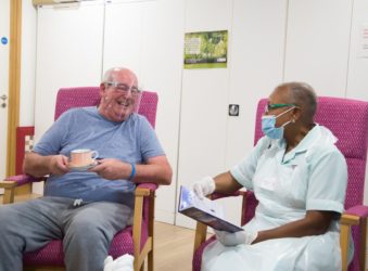 Patient wearing a perspex visor in day therapy enjoys a cup of tea, as he chats to a nurse wearing a face mask