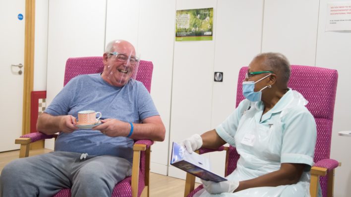Patient wearing a perspex visor in day therapy enjoys a cup of tea, as he chats to a nurse wearing a face mask