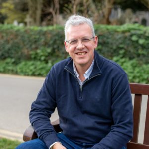 Andrew Kellard smiling, sat on a wooden bench with greenery behind him