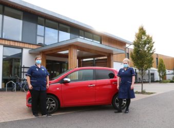 Two nurses wearing dark blue uniforms and masks, either side of a red car outside the entrance of the Hospice