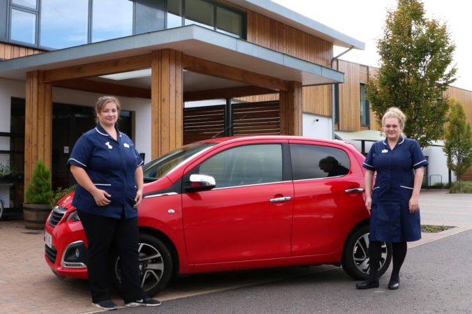 Two nurses wearing dark blue uniforms, either side of a red car outside the entrance of the Hospice