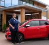 Two nurses wearing dark blue uniforms, either side of a red car outside the entrance of the Hospice