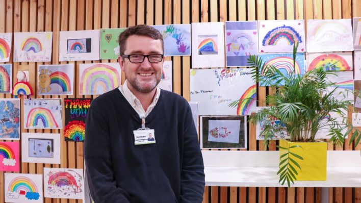 Barry Morgan, Community Team lead, smiling in front of hand-drawn rainbow pictures, in the Hospice's breakout area