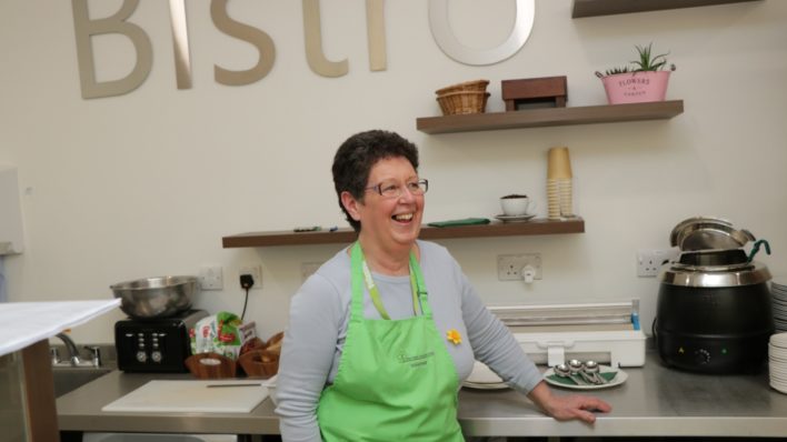 Lady volunteer standing in Bistro wearing a green apron and smiling