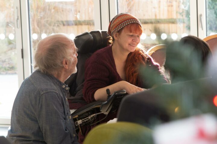 A lady with red hair and a head scarf in a wheelchair chats to a volunteer in Living Well Service lounge