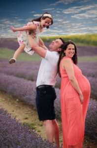 Lee holding his daughter in the air with his pregnant wife standing next to him with a lavender field behind them