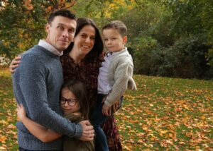 Lee with his wife and their 2 children hugging in near the Autumn leaves 