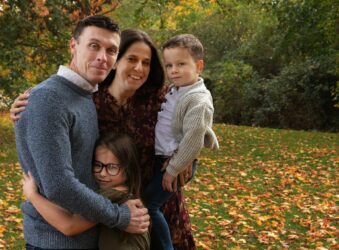 Lee with his wife and their 2 children hugging in near the Autumn leaves