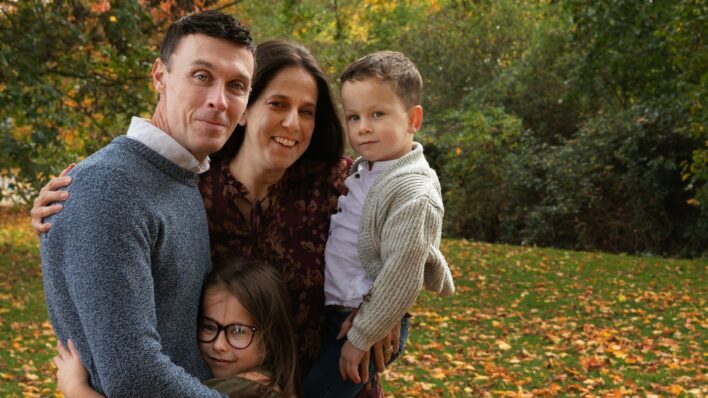 Lee with his wife and their 2 children hugging in near the Autumn leaves