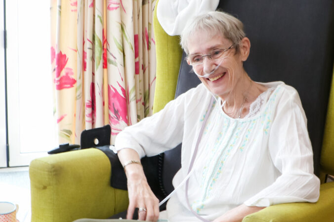 A lady with oxygen line smiles in the Living Well Lounge at the Hospice