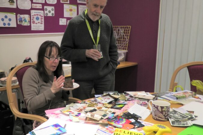 A lady holding a piece of chocolate cake explains her scrap book choices to a volunteer in the Living Well Services lounge