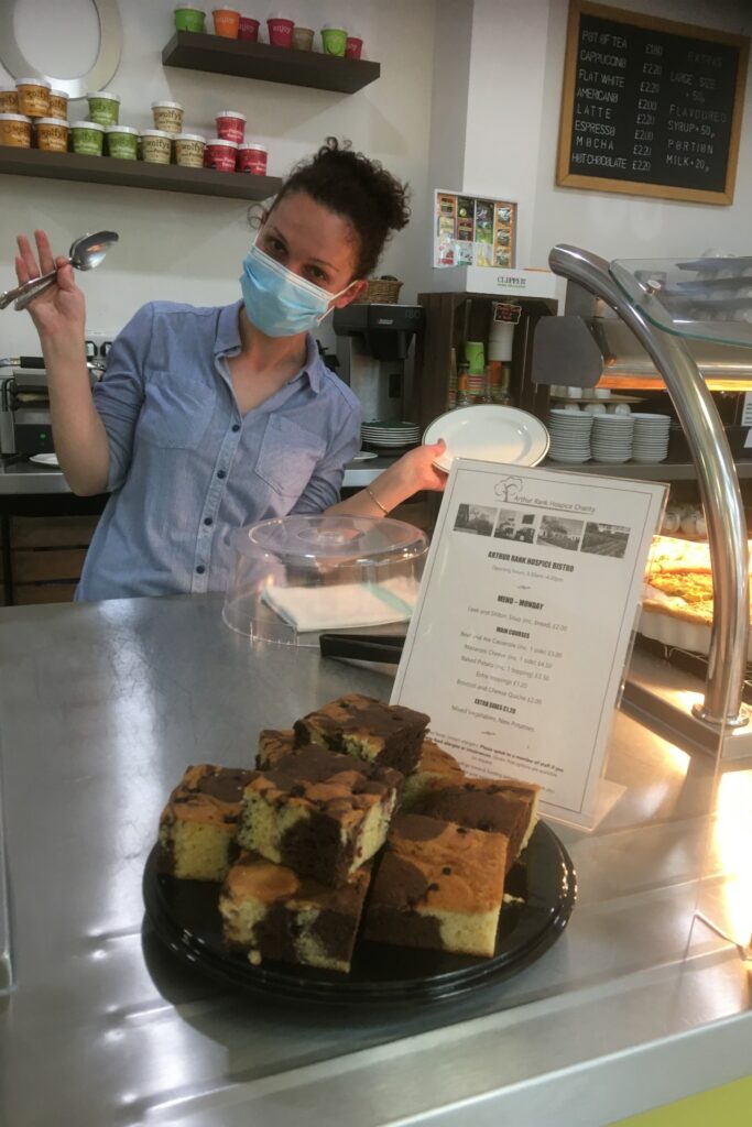 Female wearing a mask standing behind Bistro serving top, Chocolate Berry Marble Cake at the front