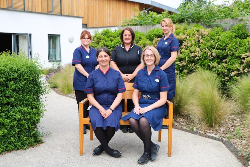 Five members of the Palliative Care Hub Team, who are Clinical Nurse Specialists in their navy uniforms, at a bench in the Hospice's gardens