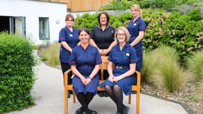 Five members of the Palliative Care Hub Team, who are Clinical Nurse Specialists in their navy uniforms, at a bench in the Hospice's gardens
