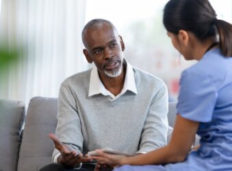 Man wearing a white shirt and grey jumper sittingon a sofa looking at a nurse wearing a blue uniform