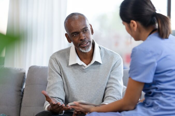 Man wearing a white shirt and grey jumper sittingon a sofa looking at a nurse wearing a blue uniform