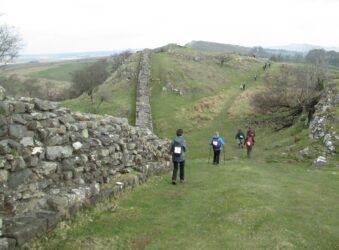 Walkers walking downhill near Hadrians Wall on the left walking towards green hills