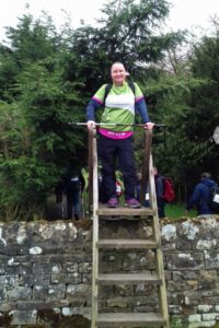 Female wearing a green, white and pink Arthur Rank Hospice Charity t-shirt at the top of a ladder at Hadrian's wall