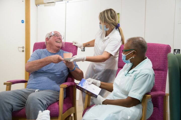 Man wearing visor smiling to a nurse with a cup of tea and another nurse sitting on a chair next to them holding an open book
