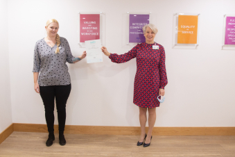 2 females hold one edge of a certificate each standing in front of framed values