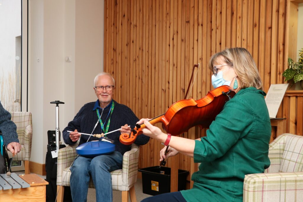 A patients enjoys playing an instrument whilst smiling at the music therapist, who is playing the viola