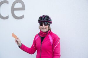 Female cyclist wearing pink jacket and black cycle helmet and glasses hold gold leaf outside white wall