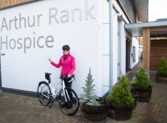 Female cyclist wearing pink and black cycling gear stands near while bike outside Arthur Rank Hospice Charity sign