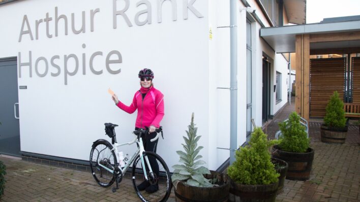 Female cyclist wearing pink and black cycling gear stands near while bike outside Arthur Rank Hospice Charity sign
