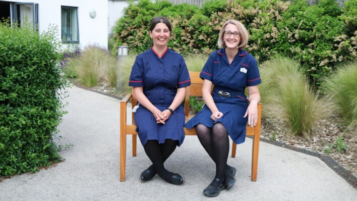 2 female nurses in blue uniforms sitting on a bench