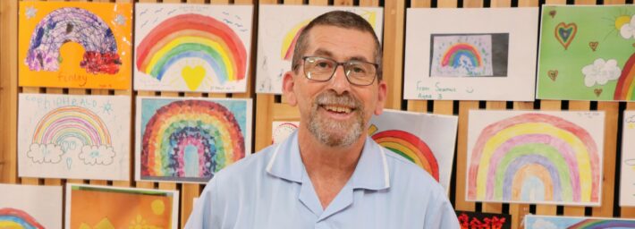 Male with glasses wearing a blue nurse uniform smiling at the camera