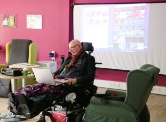 Male in a wheelchair looking at a laptop with a screen behind
