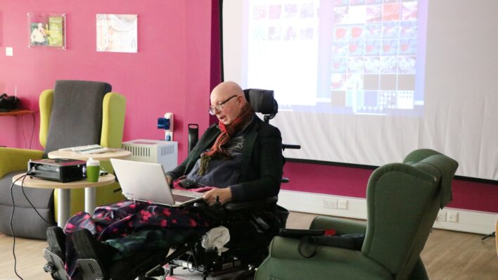 Male in a wheelchair looking at a laptop with a screen behind