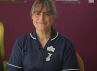 Female wearing a dark blue nurse uniform sitting in front of a pink wall