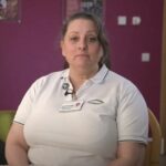 Female in a white uniform looking at the camera siting on a chair in front of a pink wall