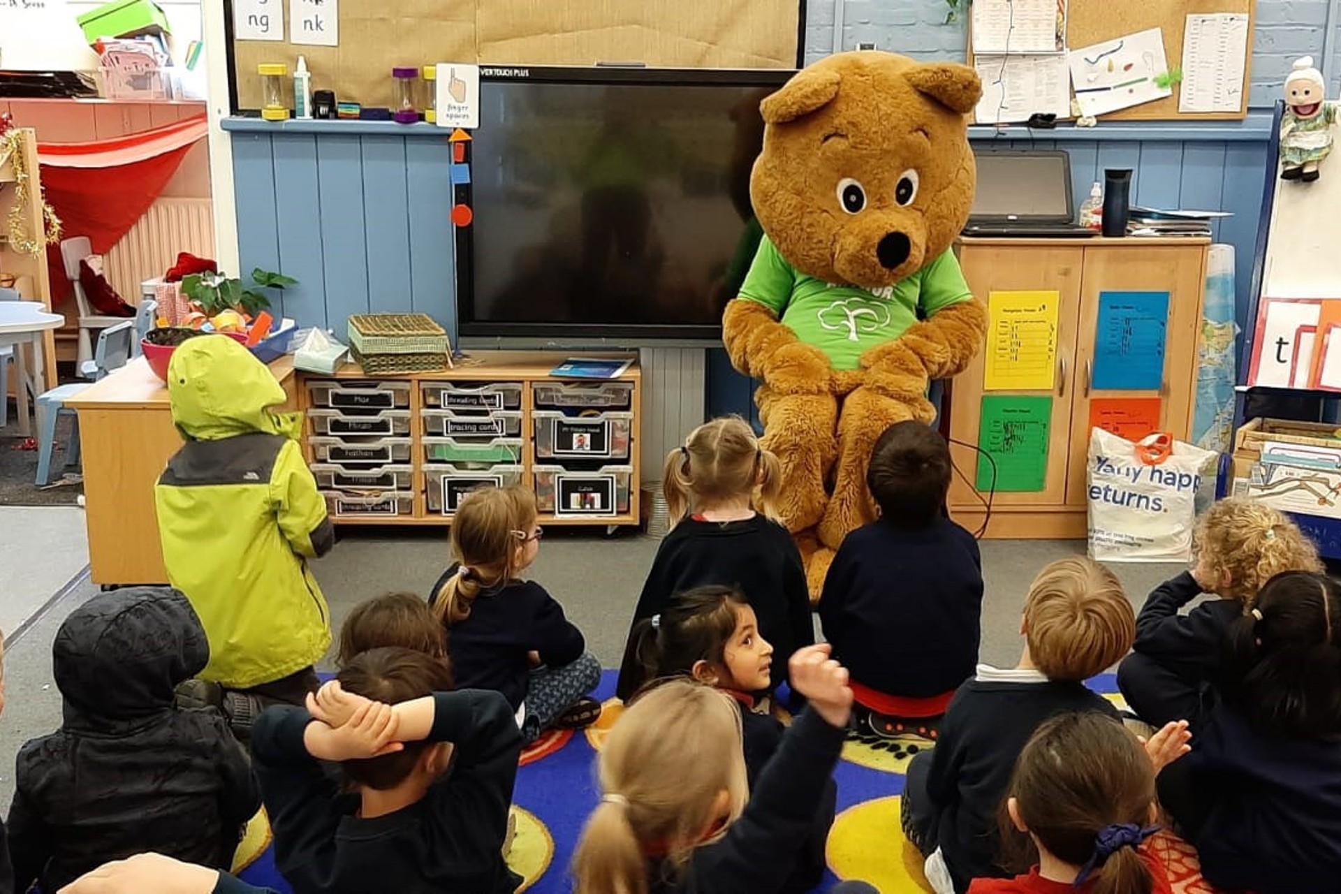 Life sized bear sitting on a chair in classroom talking to a group of children 