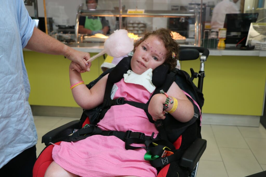 Girl in a wheelchair wearing a pink dress with a nurse holding candy floss near her face 