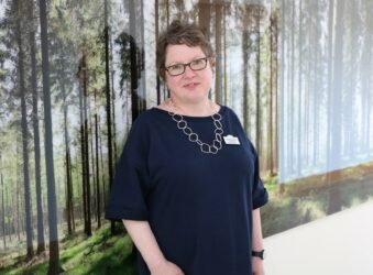 Female with glasses standing in front of a woodland picture