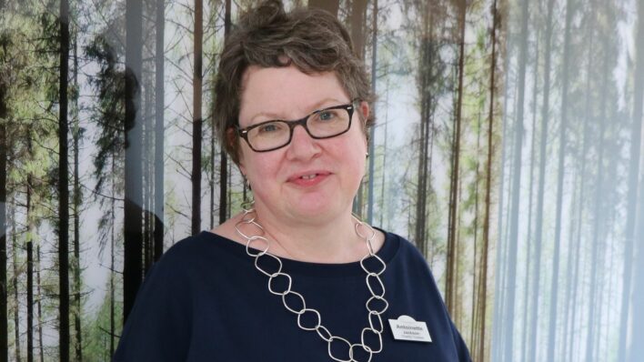 Female weraing glasses standing in front of a large phot of woodlands, looking at the camera