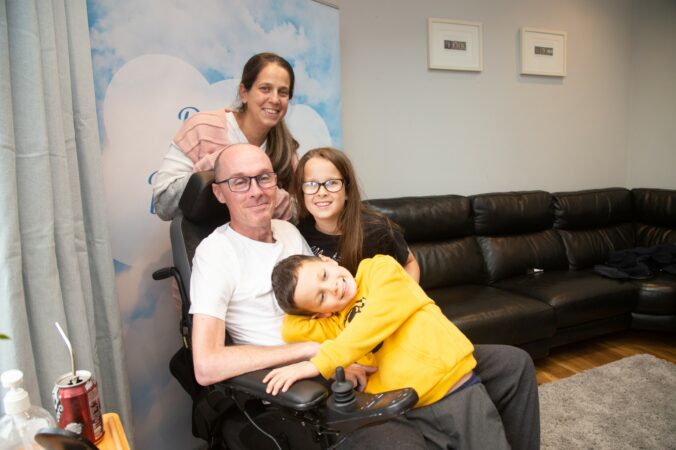 Male in a wheelchair with female adult behind him and female and male child cuddling him