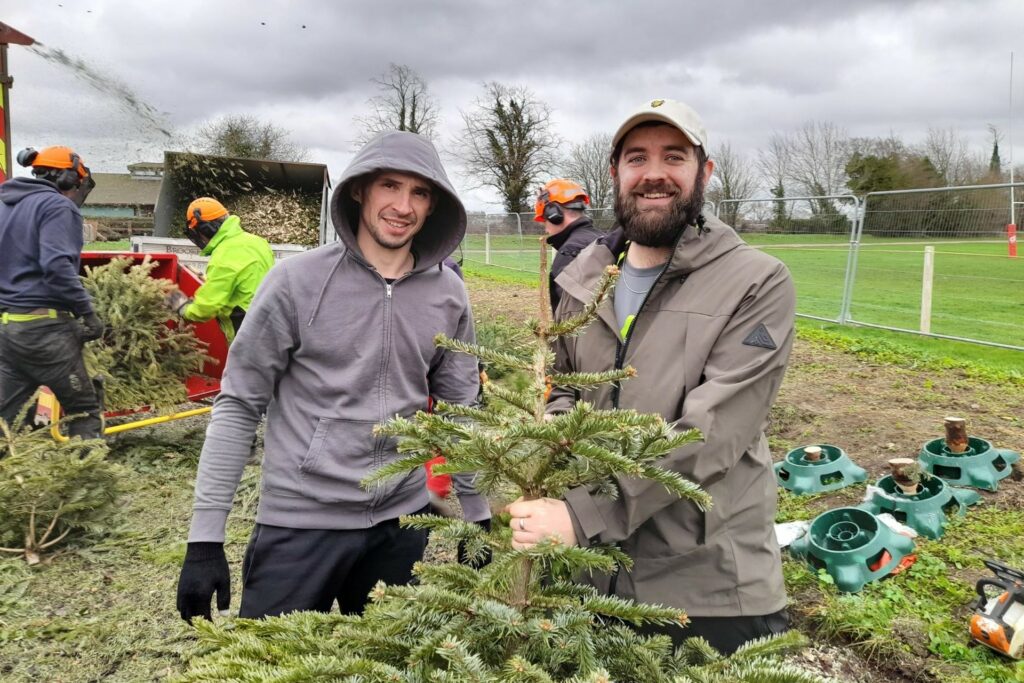 2 males wearing grey in front of a chipper with tree