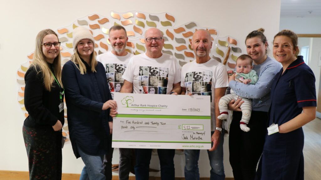 Cheque being held by 3 men with 4 women one holding a baby one in nurse uniform in front of memory tree