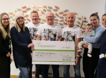 Cheque being held by 3 men with 4 women one holding a baby one in nurse uniform in front of memory tree